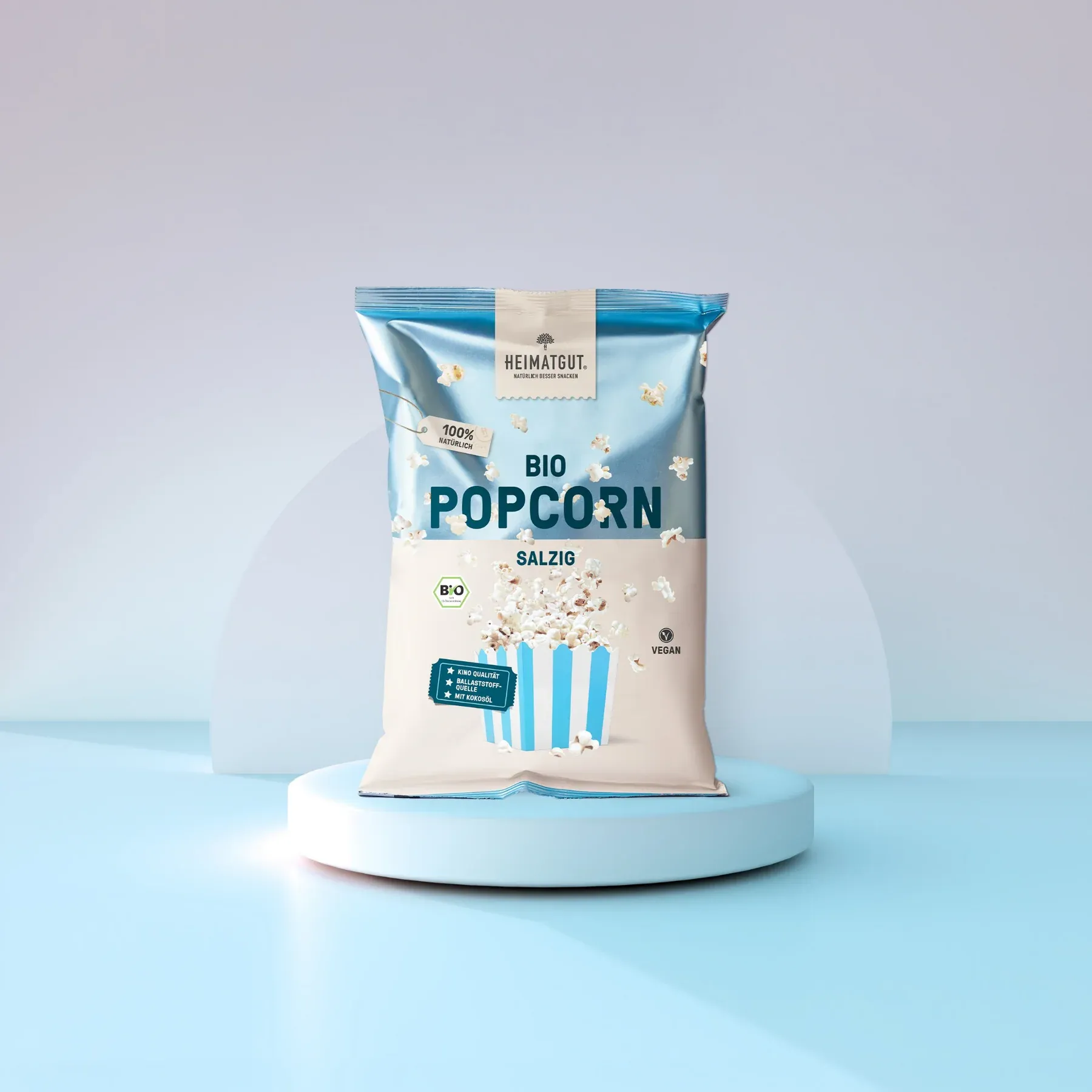 Tüte mit biologischem, salzigem Popcorn der Marke Heimatgut auf einem hellblauen Podest.