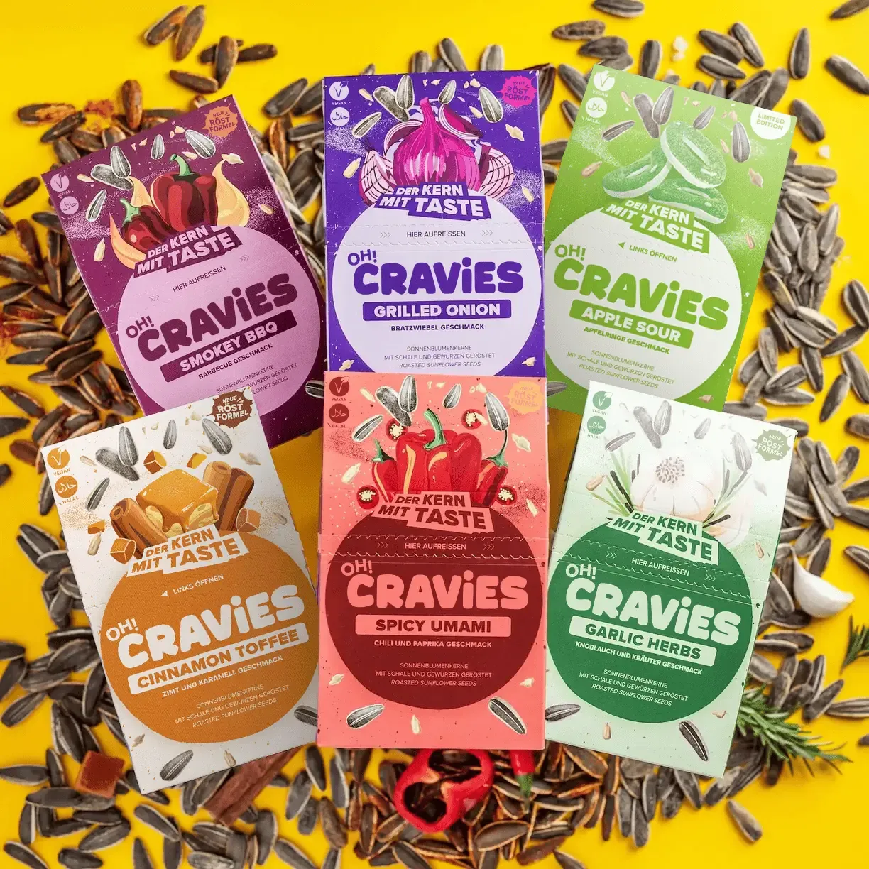 Sechs bunte Verpackungen von OH!CRAVIES sonnengetrockneten Sonnenblumenkernen in verschiedenen Geschmacksrichtungen auf gelbem Hintergrund mit gestreuten Sonnenblumenkernen.
