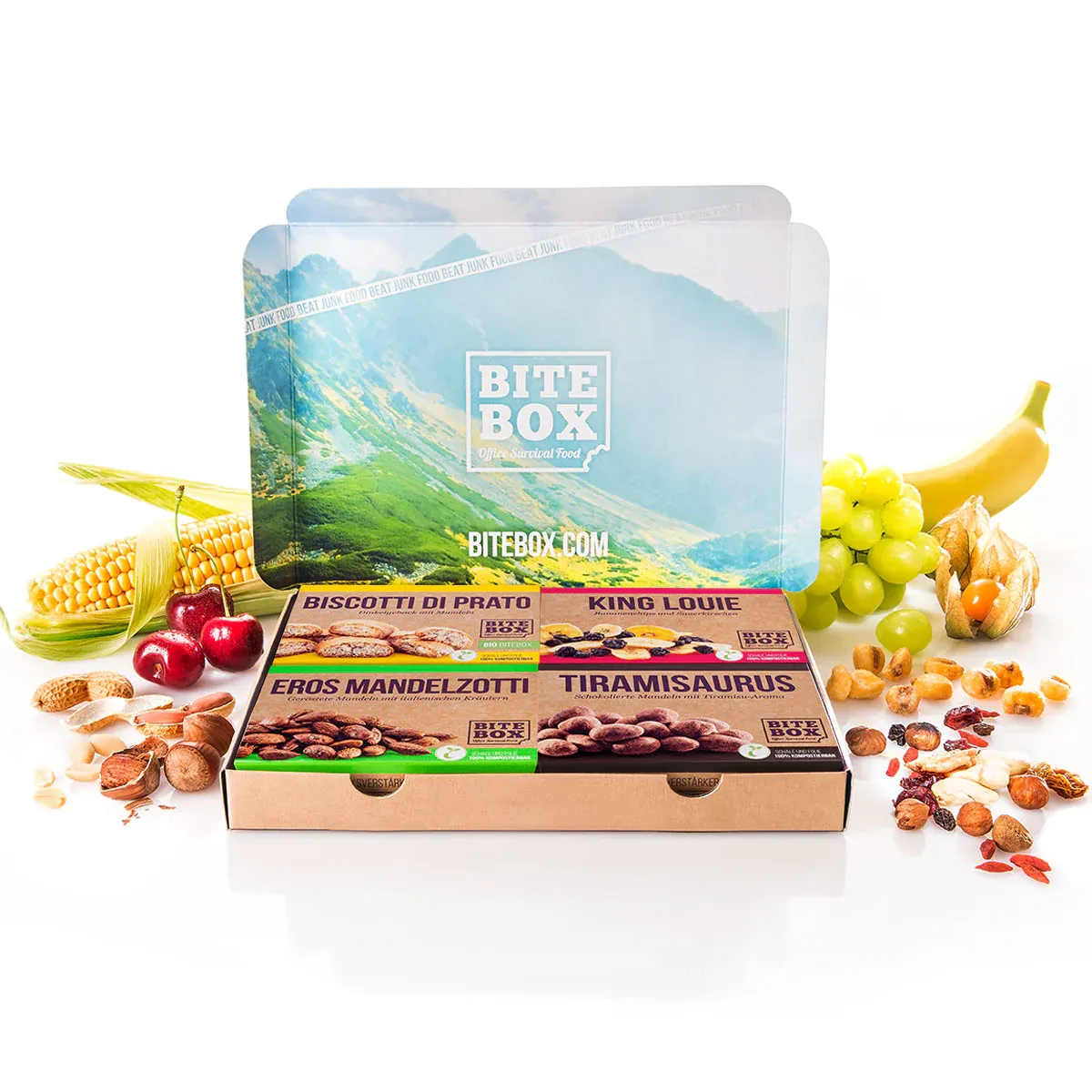 BiteBox Snack-Set mit vier Packungen Mandelsnacks auf weißem Hintergrund, umgeben von Kirschen, Mais, Nüssen, Trauben und einer Banane.