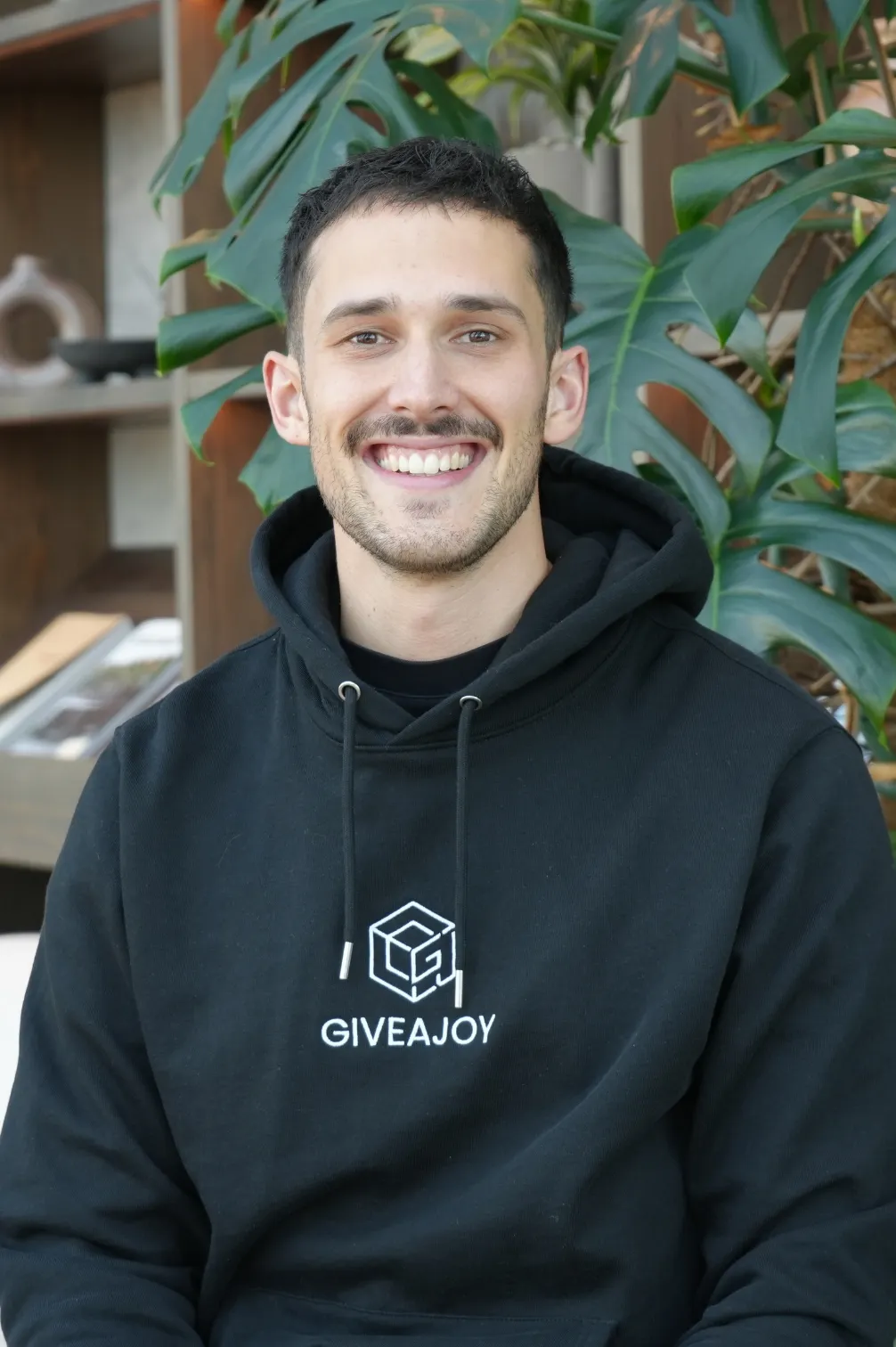 Junger Mann mit kurzem Haar und Hoodie mit Aufschrift GIVEAJOY lächelt vor grünen Pflanzen.
