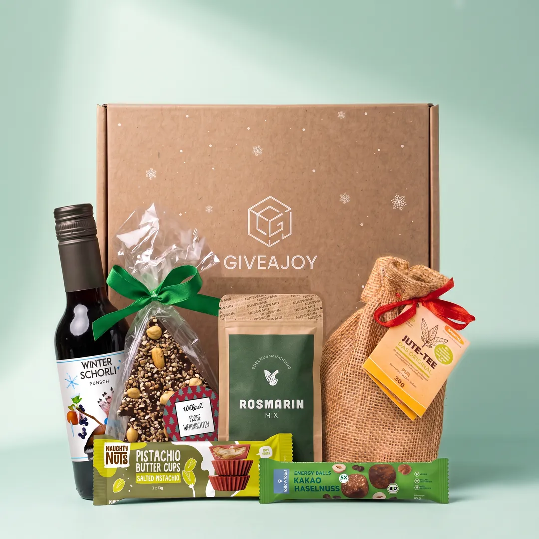 Inhalt der Weihnachtsknistern-Box vor dem GIVEAJOY-Karton: Jute-Tee, Rosmarin-Nussmix, Schokoladen-Tannenbaum, Energy Balls, alkoholfreiem Winter Punsch und Pistachio Butter Cups.