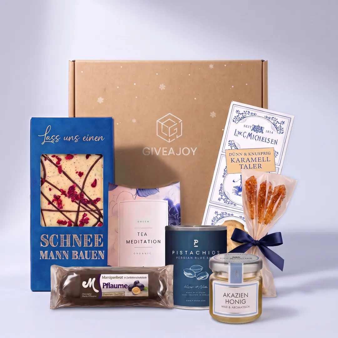Inhalt der Weihnachtszauber-Box vor dem GIVEAJOY-Karton: Kandissticks, Akazienhonig, Karamelltaler, Schokolade, Tee, Pistazien und Marzipanbrot.