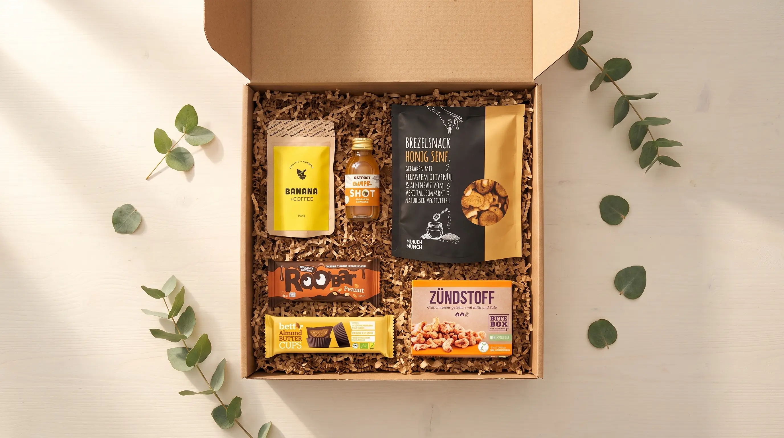 Offene Geschenkbox mit Banana-Coffee-Pulver, Ingwer-Shot, Brezelsnack Honig-Senf, Roobar Peanut, Almond Butter Cups und Zündstoff-Nüssen zwischen Eukalyptuszweigen.